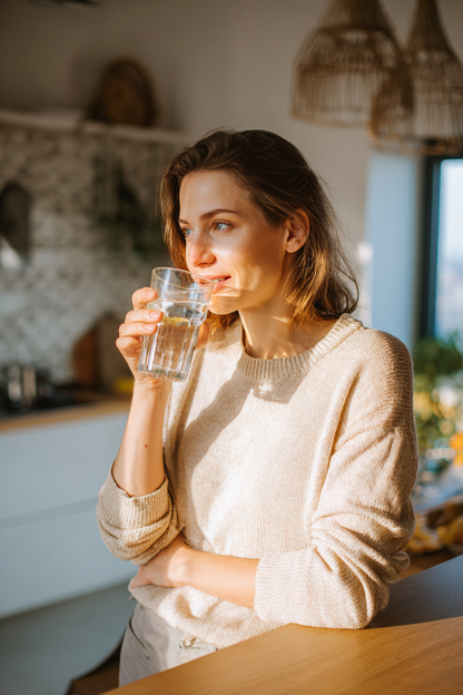 Mujer bebiendo un vaso de agua en una cocina luminosa, mostrando un momento cotidiano de toma de KLOTHOS